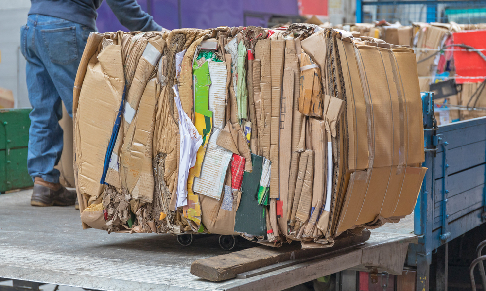 Large cardboard bale sitting on the back of a truck with other bales as a man sorts them inside a warehouse.