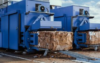 Two large blue waste compactors side by side ejecting large bales of cardboard outdoors with a blue sky and some clouds.