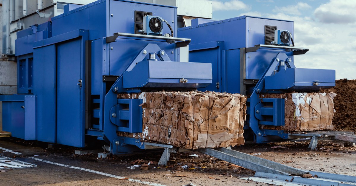 Two large blue waste compactors side by side ejecting large bales of cardboard outdoors with a blue sky and some clouds.