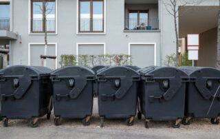 Various medium-sized black waste containers with wheels sit outside next to skinny trees in front of a residential building.