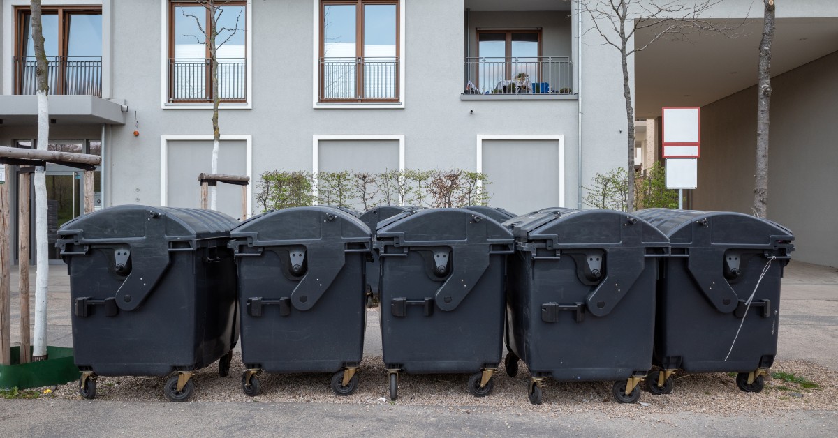 Various medium-sized black waste containers with wheels sit outside next to skinny trees in front of a residential building.