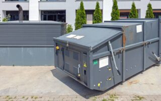 Large grey waste compactor over concrete outside a residential building with small trees behind it on a sunny day.