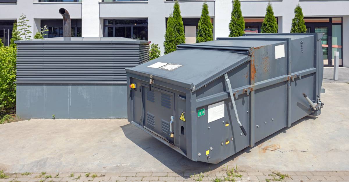 Large grey waste compactor over concrete outside a residential building with small trees behind it on a sunny day.