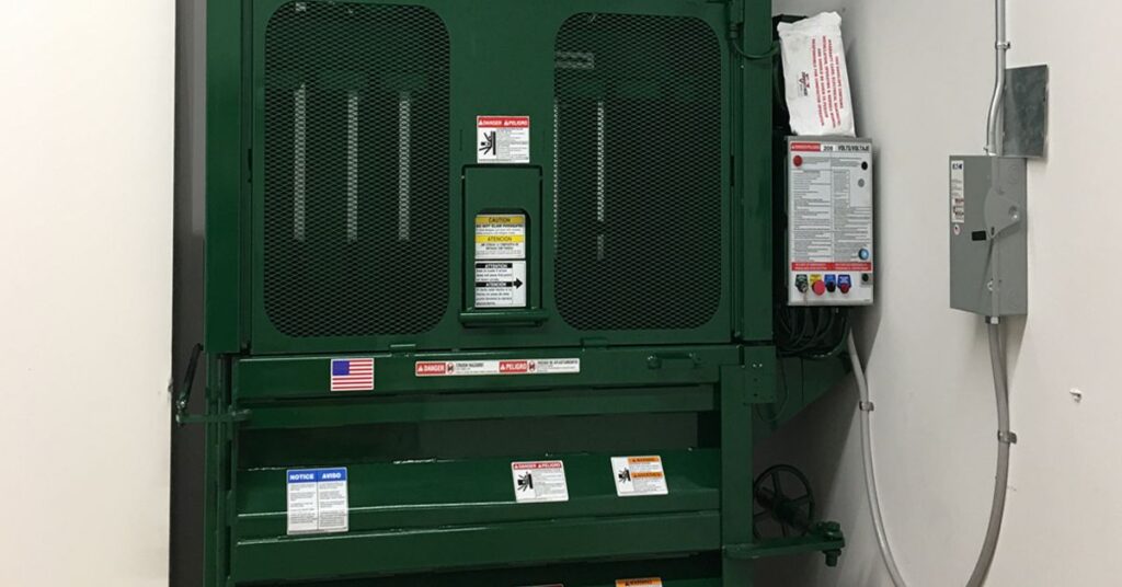 Green industrial, vertical baler in a utility room with safety labels and a control panel.