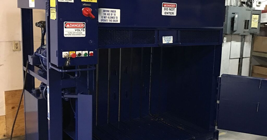 Blue industrial, vertical baler with an open loading chamber, control buttons, and safety warning sign.