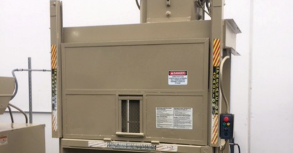 Beige vertical baler with hydraulic press enclosure, safety labels, and push-button controls in a warehouse setting.