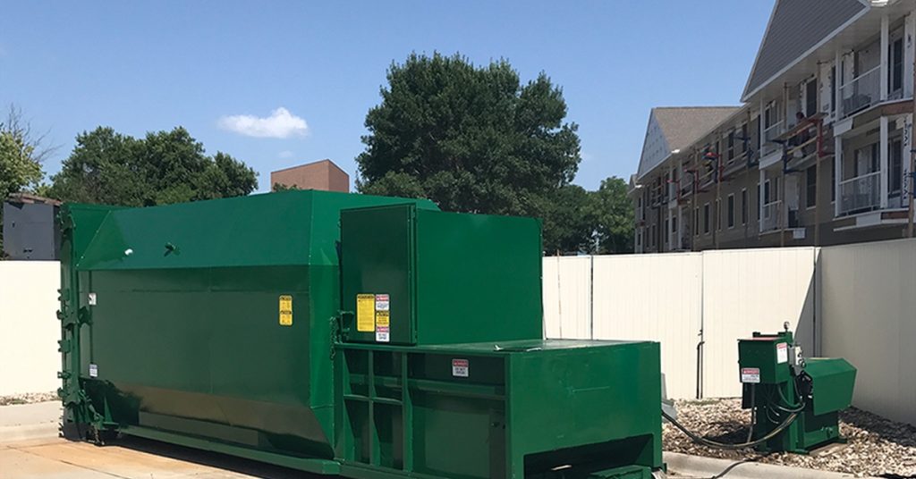 A large green self-contained compactor sits near a white fence at an apartment complex. There's units behind the fence.