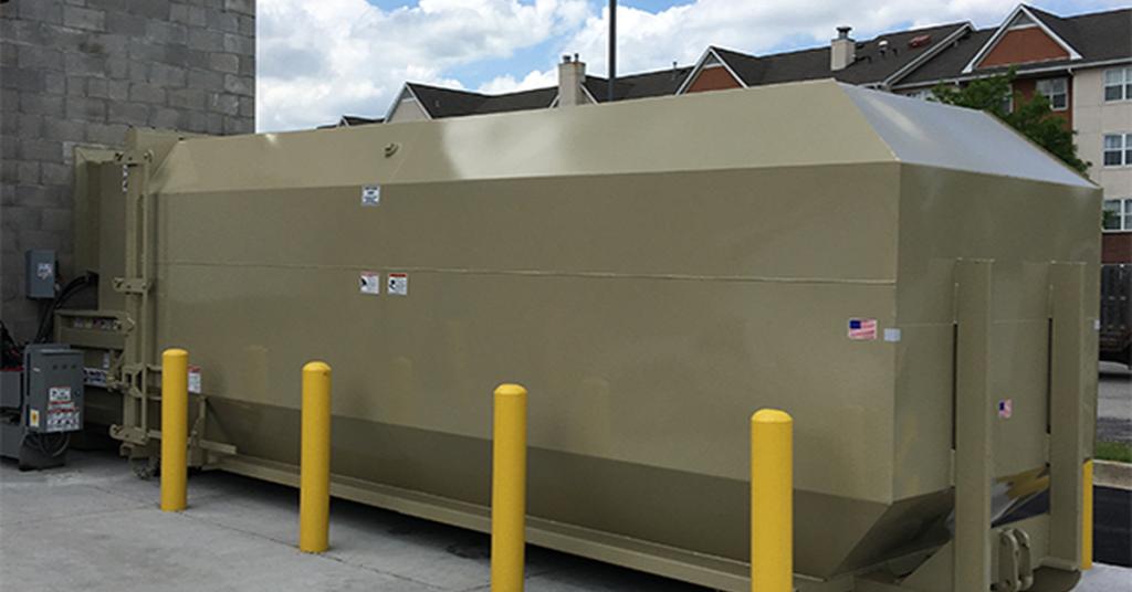 A large brown commercial compactor sits between yellow parking posts at the back corner of a building.