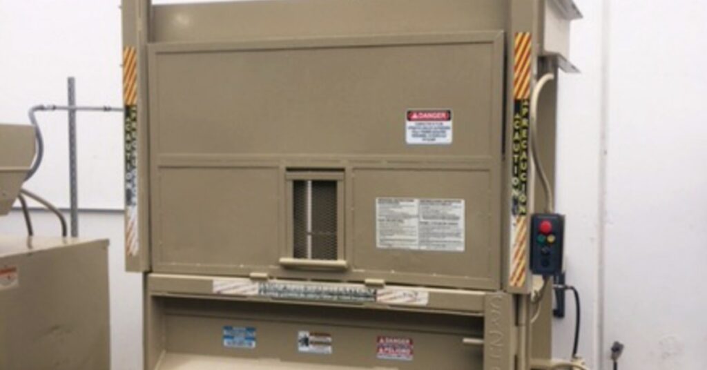 A tan industrial baler with a closed front panel, warning labels, side controls, and a wall-mounted power cord.