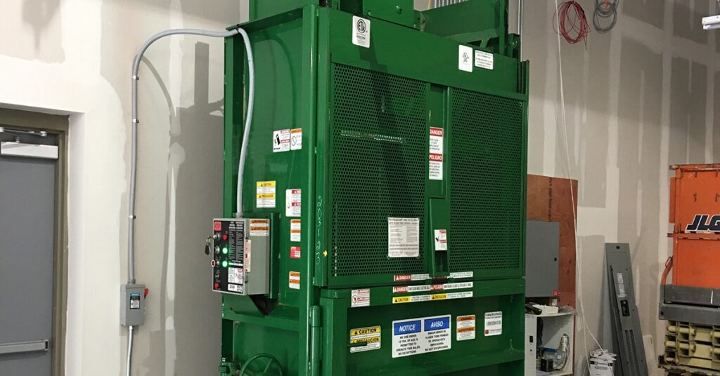 A green industrial baler with mesh front doors, warning labels, control panel, and unfinished warehouse walls.