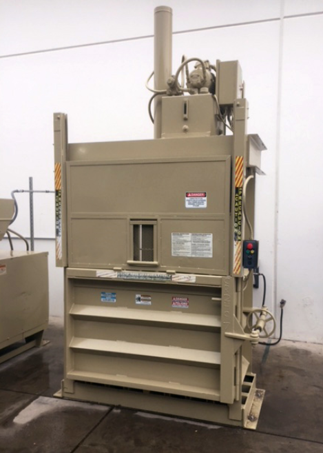 A brown, vertical baler sits at the back of a white business building. The baler is plugged in and the ground is wet.