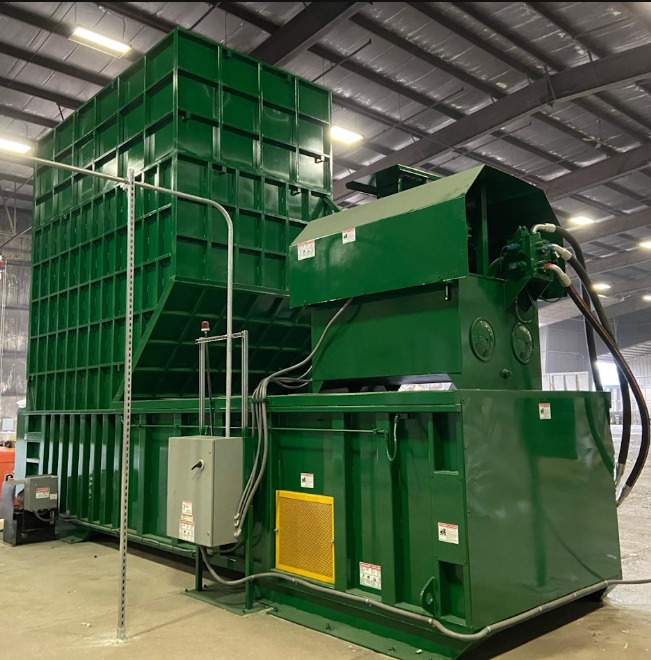 A large green self-contained baler in a commercial warehouse building. There's a silver box with plugs on the outside.
