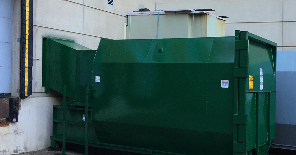 A medium-sized green self-contained compactor in the corner of a brick business. There's a loading dock to the left.