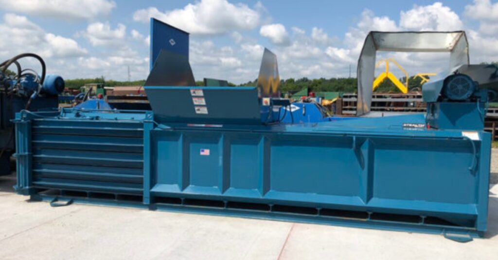 A big blue horizontal baler sits alone in the back parking lot of a commercial business. The sun shines on the baler.