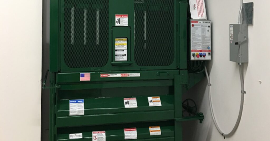 A smaller, green vertical trash compactor tucked into a nook in a warehouse's storage area. It's plugged into the wall.