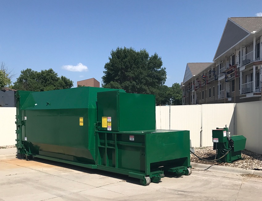 A large self-contained trash compactor at the back of a business. It's sunny outside and trees are nearby.