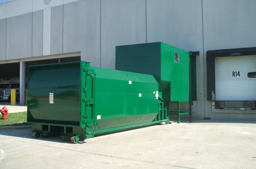 A large green self-contained trash compactor near the dock of a business warehouse. It's sunny outside.