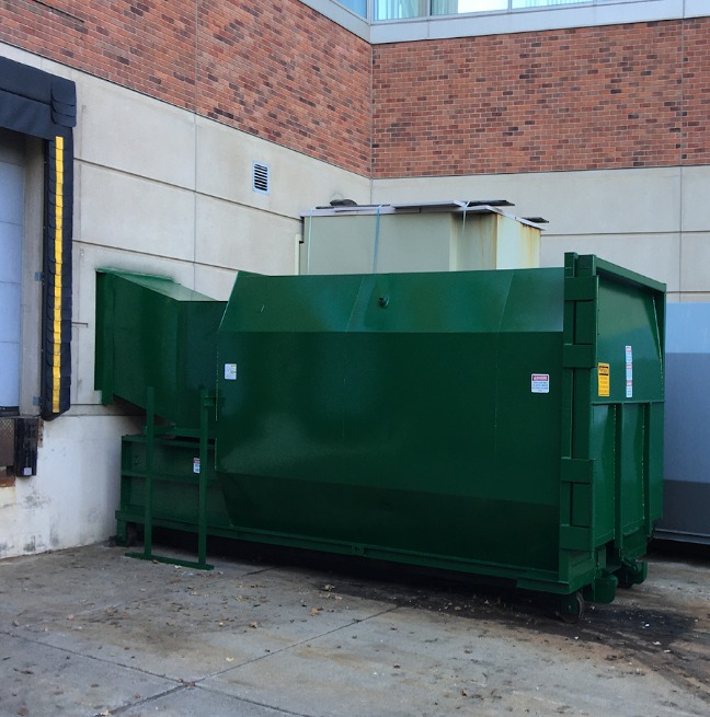 A medium-sized stationary trash compactor parked near the dock of a business facility. The building is brick.